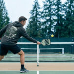 Player hitting a third shot drive from the baseline on an outdoor PNW pickleball court.